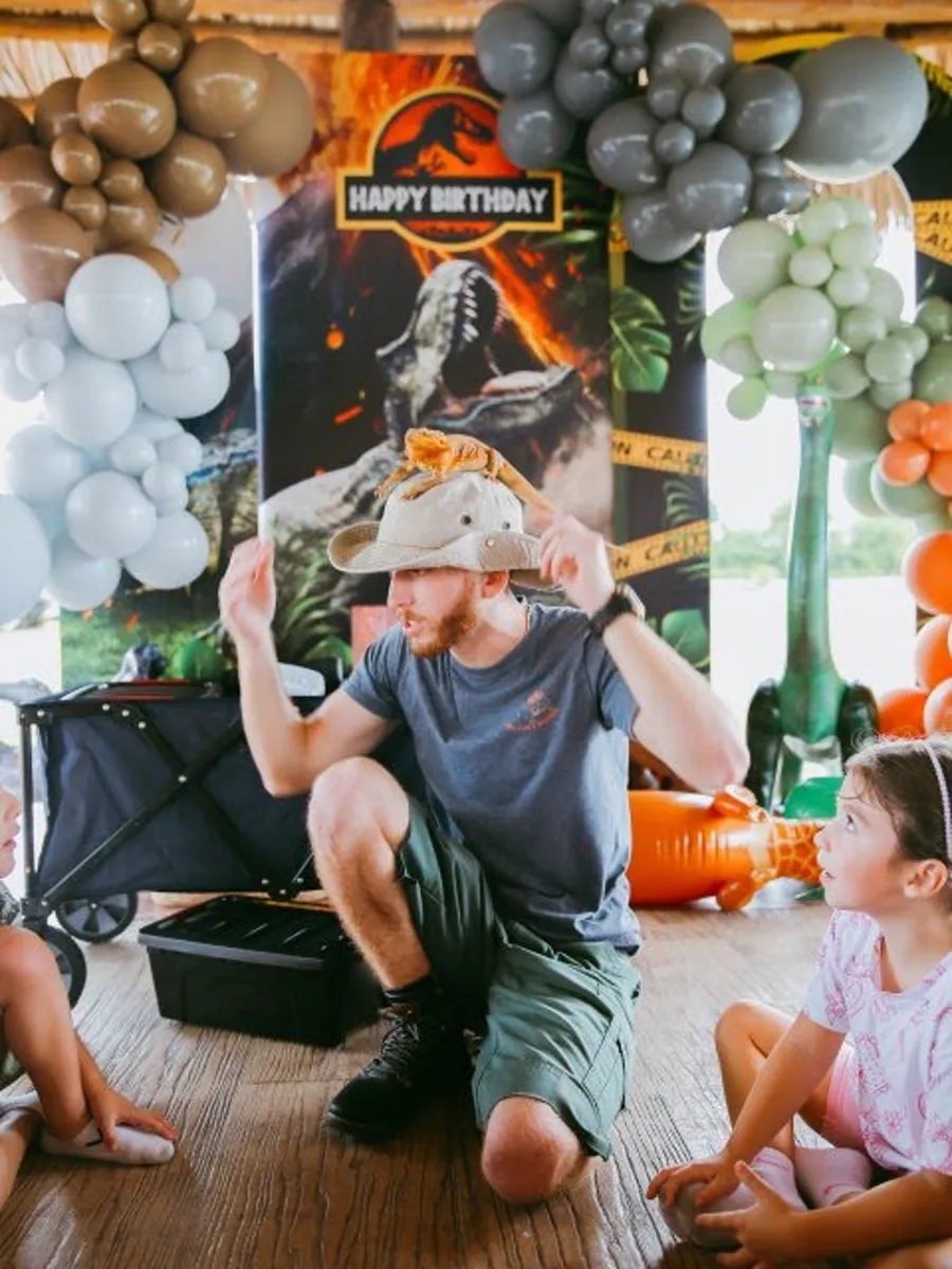 Bearded dragon riding on the handler's safari hat while kids watch at a dinosaur-themed birthday party