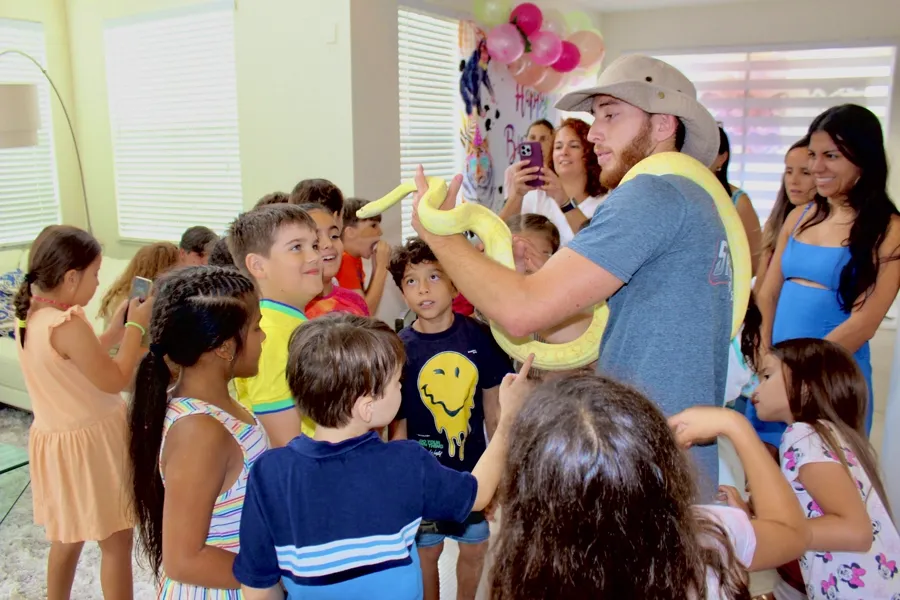 Handler carrying an albino python through a crowd of excited children at a birthday party with balloons
