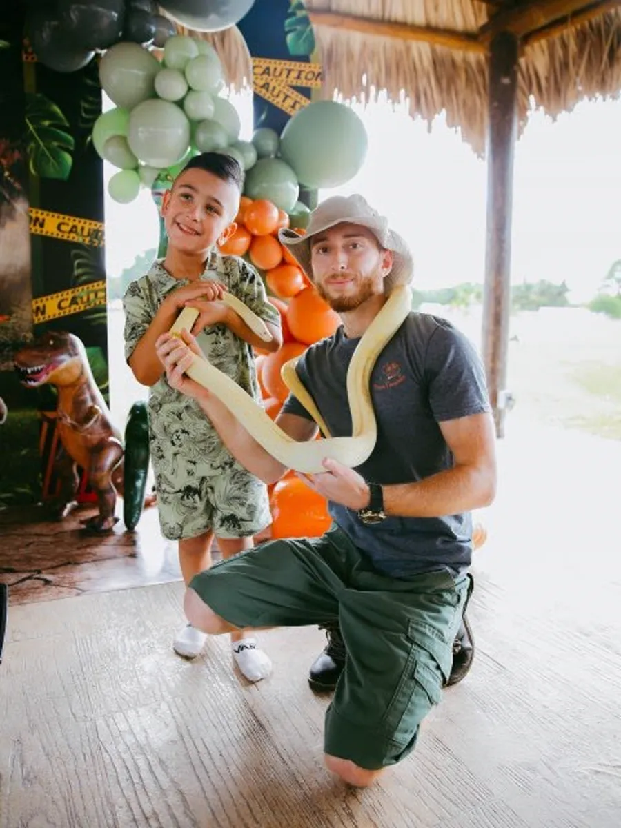 Handler kneeling beside a smiling boy with an albino python draped over his shoulders at a jungle-themed birthday party