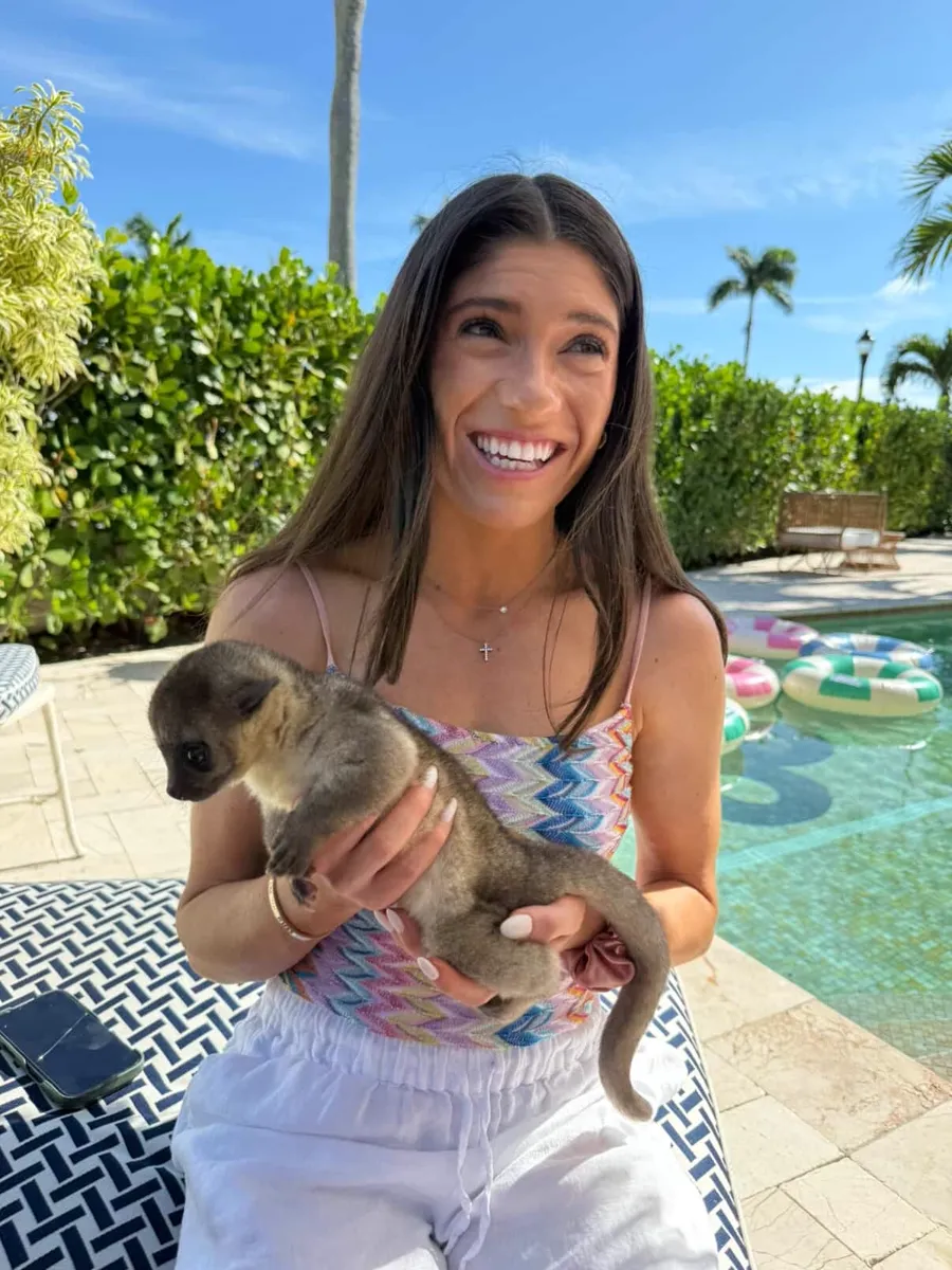 Laughing woman holding a baby kinkajou beside a pool with palm trees behind